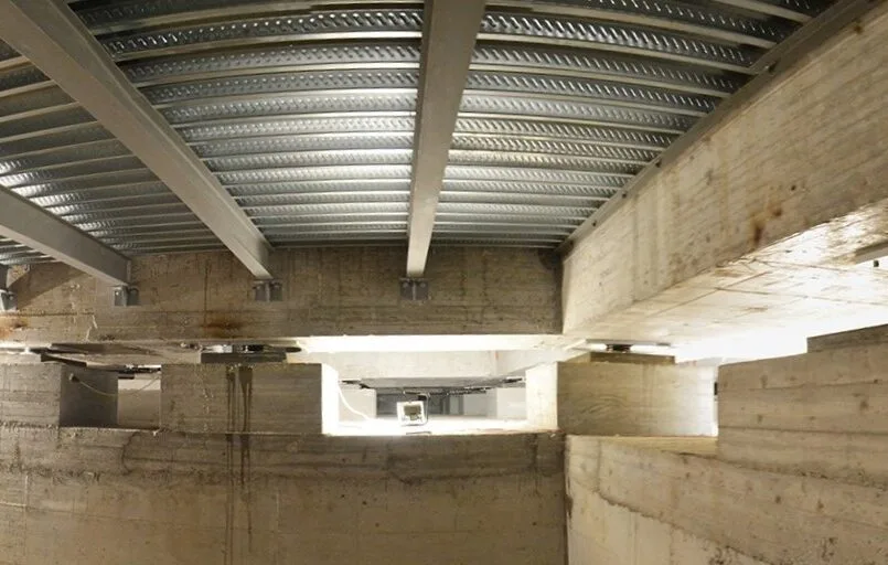 Isolatori sismici neri tra plinti in calcestruzzo, travi e lamiera grecata viste dal vano fondazioni di un edificio