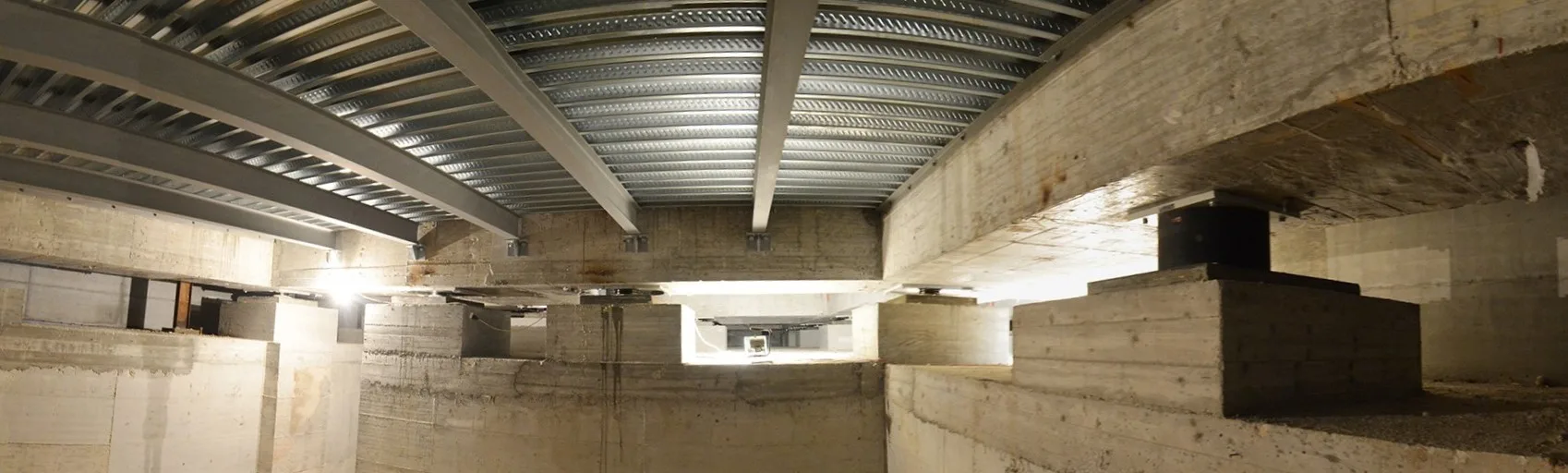 Isolatori sismici neri tra plinti in calcestruzzo, travi e lamiera grecata viste dal vano fondazioni di un edificio