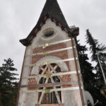 Torre in mattoni e pietra rinforzata con fasce bianche e travature lignee, cielo grigio e alberi ai lati del cantiere