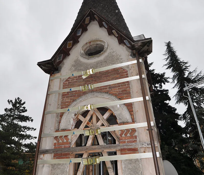 Torre in mattoni e pietra rinforzata con fasce bianche e travature lignee, cielo grigio e alberi ai lati del cantiere