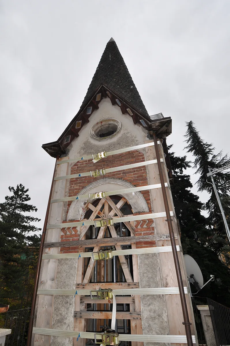 Torre in mattoni e pietra rinforzata con fasce bianche e travature lignee, cielo grigio e alberi ai lati del cantiere