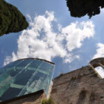 Vista dal basso verso il cielo: torre vetrata con ferramenta a ragno accanto a un alto muro in pietra e cipressi
