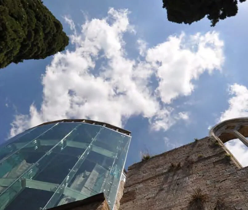 Vista dal basso verso il cielo: torre vetrata con ferramenta a ragno accanto a un alto muro in pietra e cipressi