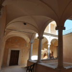 Porticato con volte in pietra chiara e colonne su muri ocra scrostati, seduta nera e cortile illuminato sullo sfondo