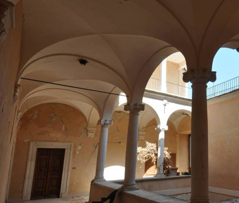 Porticato con volte in pietra chiara e colonne su muri ocra scrostati, seduta nera e cortile illuminato sullo sfondo