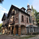 Edificio in mattoni con torre conica, loggiato ad archi e rinforzi lignei alle colonne, prato e alberi intorno