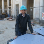 Ingegnere con casco turchese appoggiato a una lastra segnata Emiciclo, colonne classiche e recinzione del cantiere sullo sfondo
