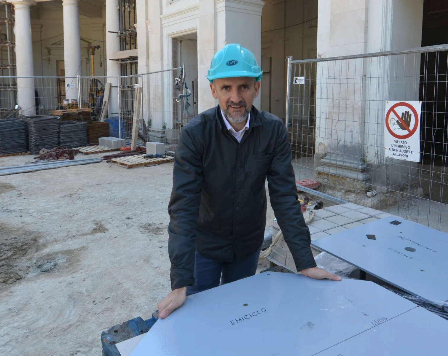 Ingegnere con casco turchese appoggiato a una lastra segnata Emiciclo, colonne classiche e recinzione del cantiere sullo sfondo